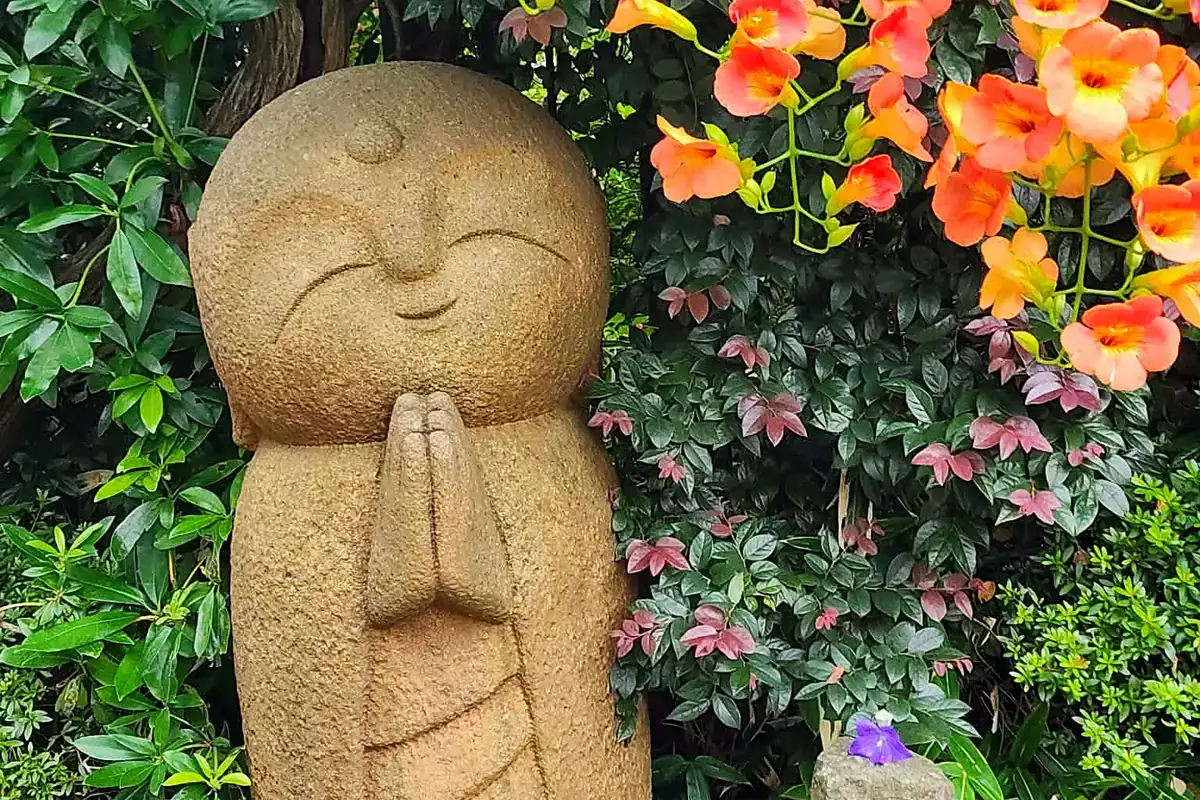 Une statue de jizō en pierre, mains jointes, entourée de fleurs orange à Hase-dera