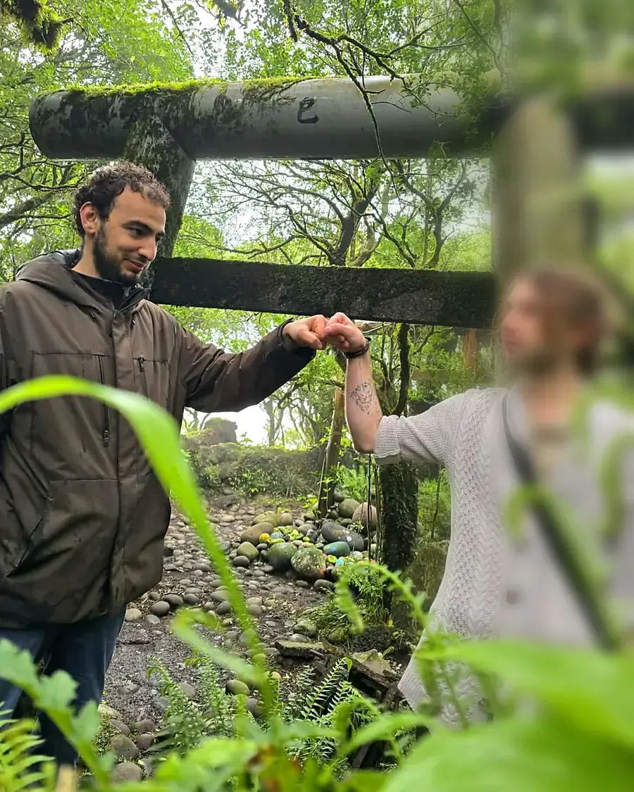 Le guide et une visiteuse se saluent sous un torii de pierre couvert de mousse, en pleine forêt